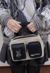 Mains tenant un sac Karl Lagerfeld Jeans beige et noir avec deux poches avant, porté sur un pull à motifs et une jupe à carreaux.