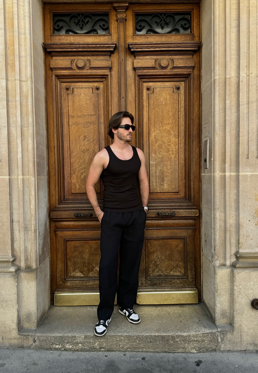 Μαύρο στενό μπλουζάκι και χαλαρές σκούρες παντελόνες φορεμένες με μαύρα και άσπρα αθλητικά παπούτσια, στέκεται δίπλα σε διακοσμητικές ξύλινες πόρτες με περίπλοκα σχέδια.