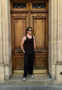 Μαύρο στενό μπλουζάκι και χαλαρές σκούρες παντελόνες φορεμένες με μαύρα και άσπρα αθλητικά παπούτσια, στέκεται δίπλα σε διακοσμητικές ξύλινες πόρτες με περίπλοκα σχέδια.