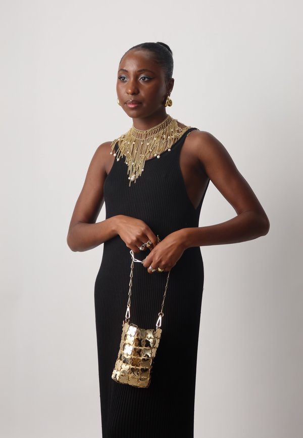 Czarna prążkowana sukienka z warstwowym złotym naszyjnikiem i wiszącymi kolczykami; trzyma małą torebkę na łańcuszku ze złotym teksturowanym wzorem.