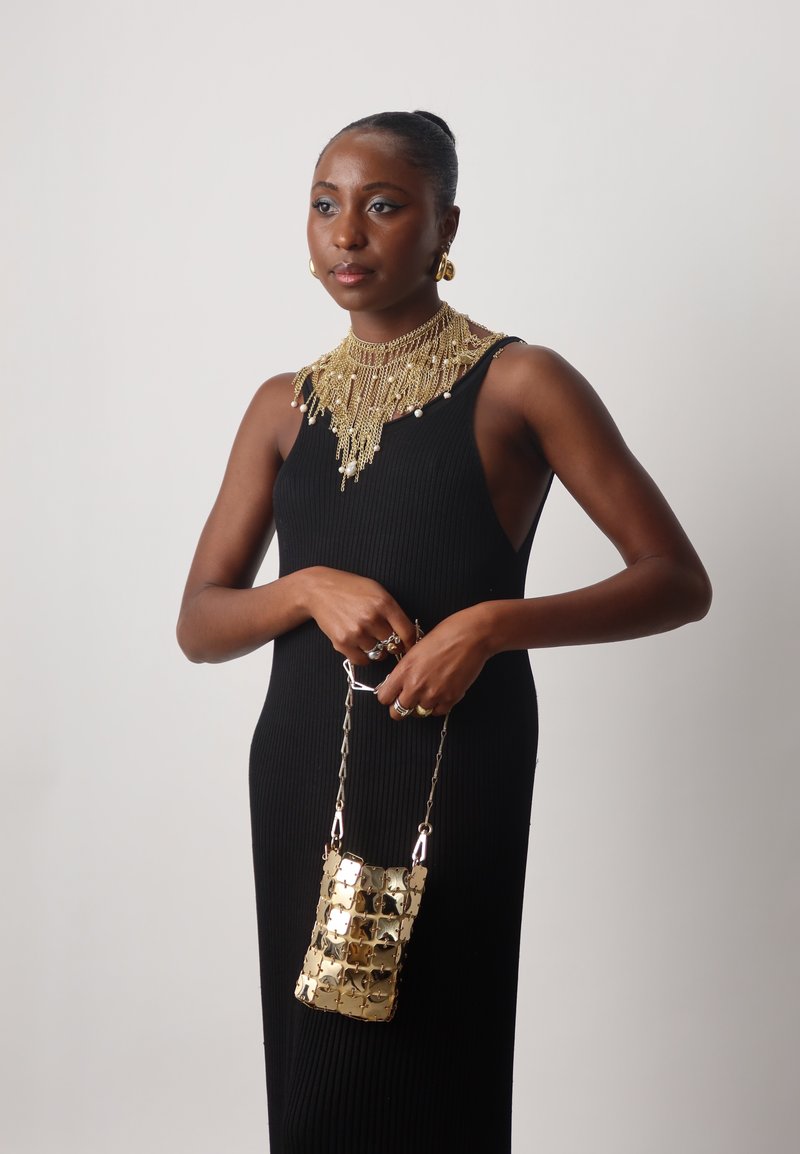 Robe noire côtelée avec un collier doré superposé et des boucles d'oreilles pendantes ; tenant un petit sac à chaîne doré avec un motif texturé.