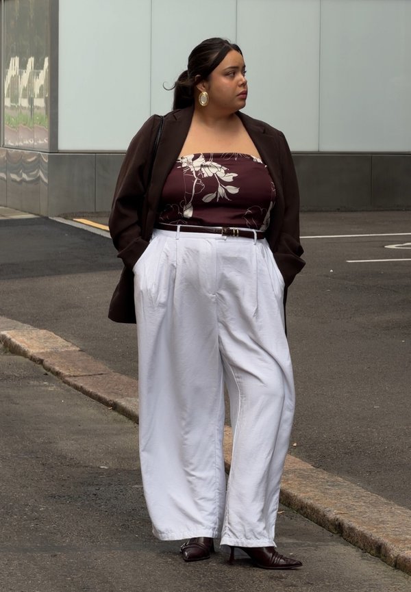 Mujer de pie en una esquina de la calle con una chaqueta marrón, top tubo floral, pantalones blancos de pierna ancha y zapatos oscuros con tacón, mirando hacia su derecha.
