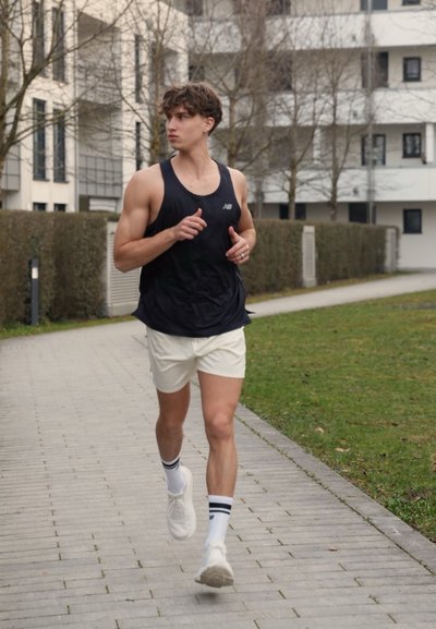 Joven corriendo en un sendero pavimentado de un parque urbano, vestido con una camiseta sin mangas negra, pantalones cortos blancos, calcetines blancos y zapatillas de correr blancas.