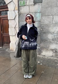 Veste noire duveteuse zippée avec un motif de cheval enneigé, associée à un pantalon cargo large vert olive et des chaussures de sport blanches. Sac gris avec des accents.