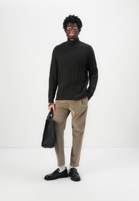 Črna pletena pulover s turtleneck ovratnikom in teksturiranim vzorcem, bež prugaste hlače, črna usnjena torba in črne slip-on čevlje z belimi nogavicami.