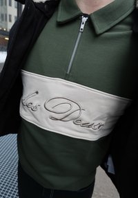 Gros plan d'une personne portant un pull vert avec une bande horizontale beige et un texte en cursive brodé sur la poitrine.