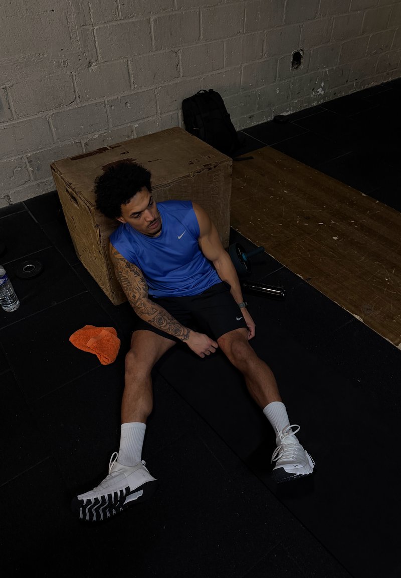 Giovane uomo con canotta blu e pantaloncini neri seduto sul pavimento della palestra, appoggiato a una cassa di legno, con bottiglia d'acqua, asciugamano e zaino nelle vicinanze.