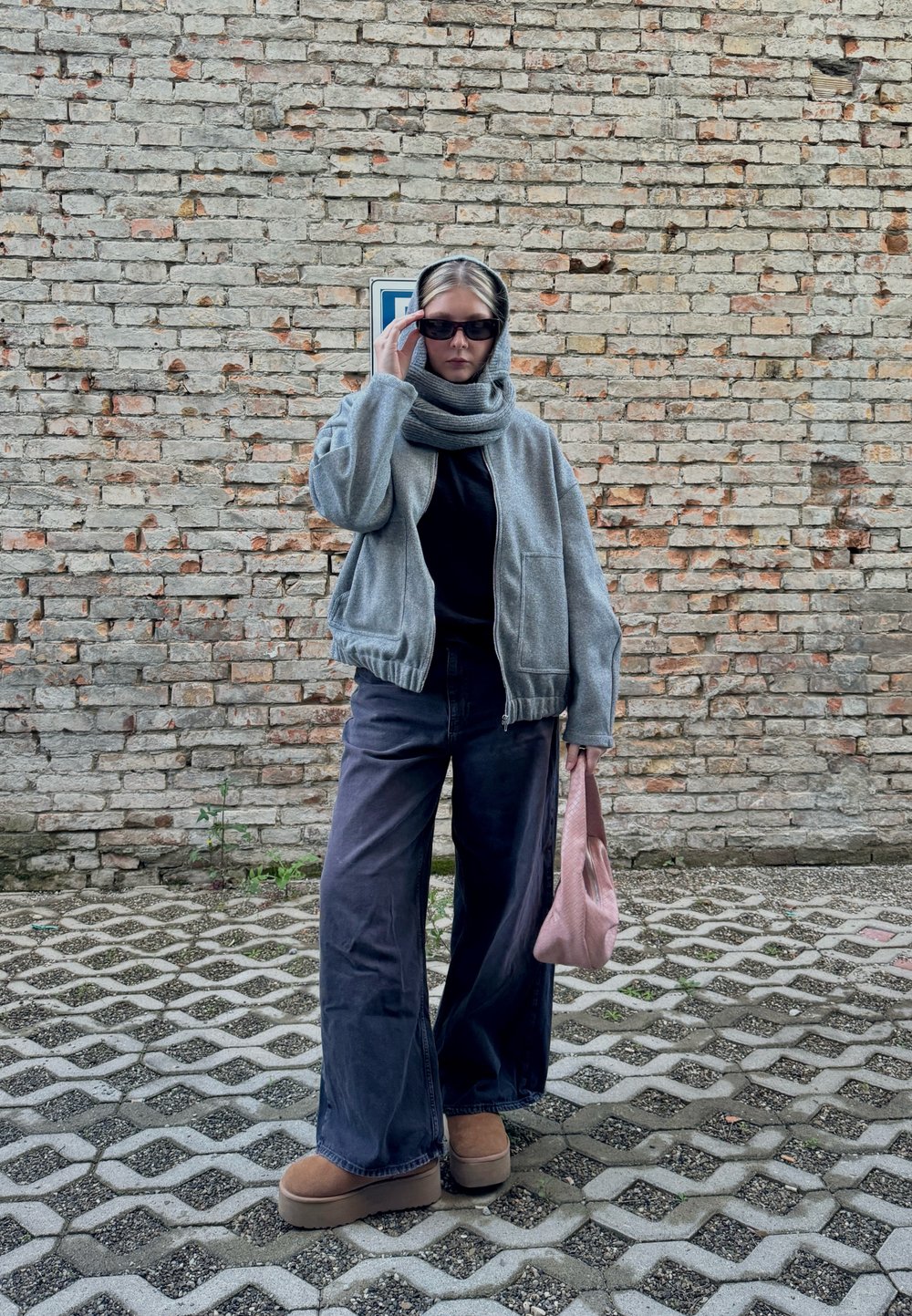 Casaco bomber cinza com fecho, blusa preta, jeans largos escuros, botas de plataforma castanhas, cachecol cinza e bolsa rosa em frente a uma parede de tijolos.