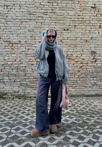 Casaco bomber cinza com fecho, blusa preta, jeans largos escuros, botas de plataforma castanhas, cachecol cinza e bolsa rosa em frente a uma parede de tijolos.