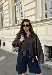 Hnědá kožená bomber bunda s mírným leskem, volným střihem, zipovým zapínáním a nastavitelným bočním popruhem. Noste ji s tmavými kraťasy a cvočkovou taškou.