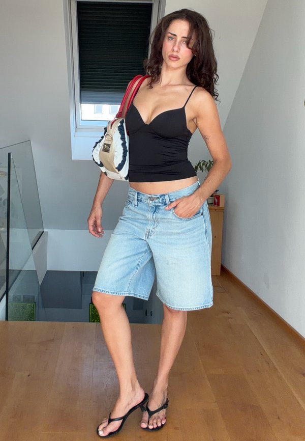 Femme debout à l'intérieur portant un top noir à fines bretelles, un short en jean longueur genoux, des sandales noires et portant un sac à main blanc et rouge à l'épaule.
