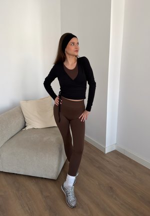 Mujer con leggings marrones, top cruzado negro y cinta en la cabeza, de pie con las manos en las caderas cerca de un sofá beige sobre suelo de madera.