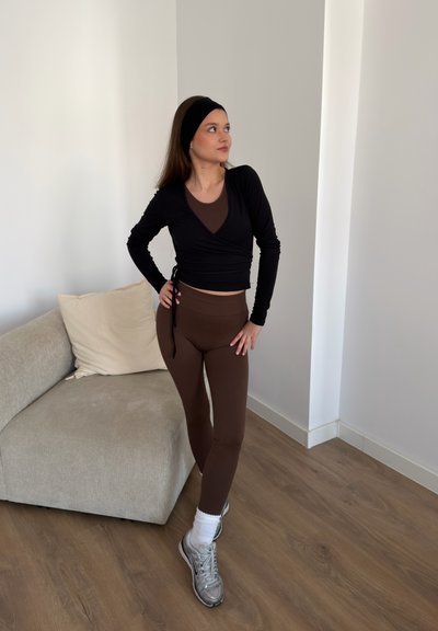 Mujer con leggings marrones, top cruzado negro y cinta en la cabeza, de pie con las manos en las caderas cerca de un sofá beige sobre suelo de madera.
