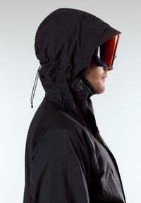 Άνδρας που φοράει μαύρο μπουφάν με κουκού λα και μεγάλα κόκκινα γυαλιά σκι, κοιτάζοντας δεξιά μπροστά σε απλό ανοιχτόχρωμο φόντο.