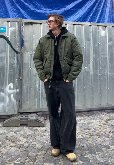 Chaqueta bomber verde sobre una sudadera negra, combinada con unos jeans negros holgados y zuecos color marrón. El fondo presenta una cerca de metal y un suelo de adoquines.