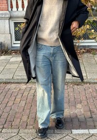 Person iført lyseblå jeans med vide ben, beige sweater, sort frakke med ternet foer og sorte sko, der står på en brostensbelagt fortov.