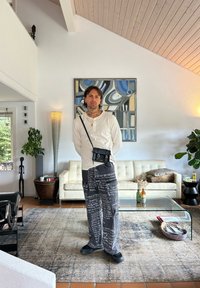 Jeune homme tatoué se tient dans le salon portant une chemise blanche à manches longues, un pantalon noir imprimé, un sac en bandoulière, près d'un canapé blanc et d'une table basse en verre.