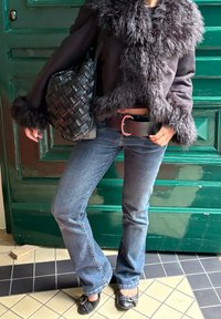 Fekete műszőrme kabát, szőtt fekete kézi táskával, kék farmer flared nadrággal és fekete lapos cipővel. Az öv fémből készült csattal rendelkezik. Geometrikus padlólapok.