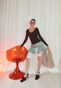 Femme en haut noir, mini-jupe en denim, bas blancs en dentelle jusqu'aux cuisses et talons, pose à côté d'une chaise translucide orange, tenant un sac beige duveteux.