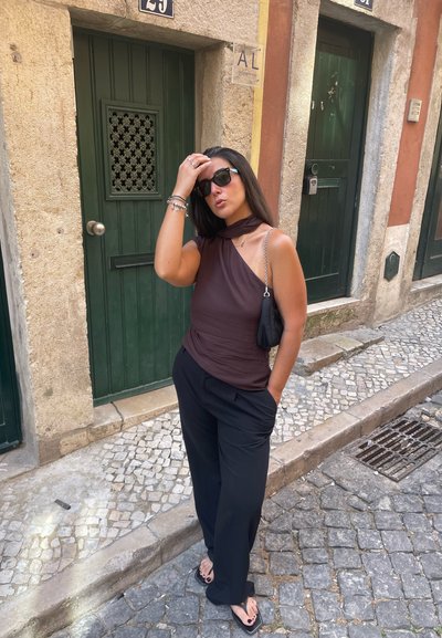 Top marrón de un solo hombro con un detalle de volantes; pantalones anchos negros; bolso negro; gafas de sol oscuras; de pie contra una pared de piedra texturizada.