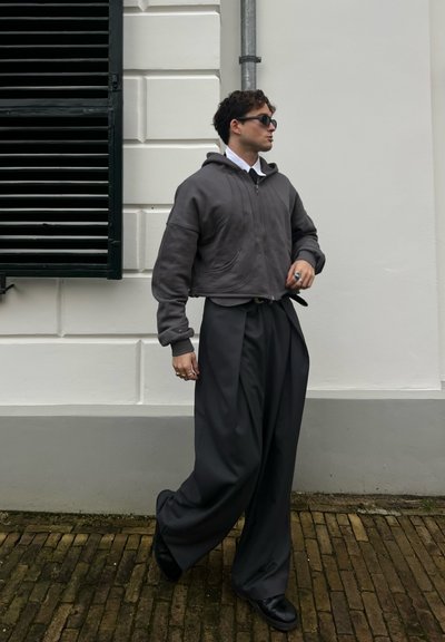 Sudadera gris recortada con cremallera frontal, combinada con pantalones oscuros de gran tamaño en gris. Zapatos negros y gafas de sol completan el atuendo.