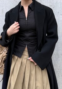 Manteau en laine noire porté sur un haut noir ajusté avec une fermeture éclair, associé à une jupe plissée beige. La tenue est accessoirisée d'un sac marron clair.
