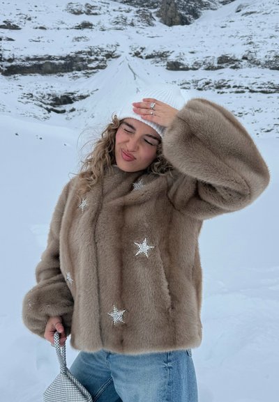Mujer con abrigo de piel color marrón claro con adornos de estrellas y gorro blanco de punto, sosteniendo una pequeña bolsa, de pie en un paisaje montañoso nevado.