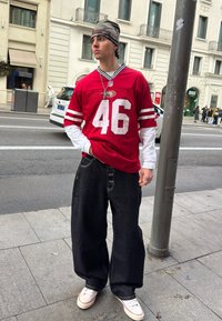 Jeune homme portant un maillot NFL rouge numéro 46, un bonnet camouflage, un jean large noir et des baskets blanches, debout sur un trottoir en ville.