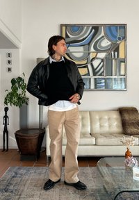 Veste en cuir noire sur un pull noir, chemise blanche à col, pantalon beige et chaussures noires. Debout dans une pièce avec de l'art abstrait et des meubles.
