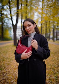 Bolsa de mão de couro vermelho, com um estilo hobo, apresentando uma forma arredondada e uma alça curta, segurada contra um fundo de folhas de outono.