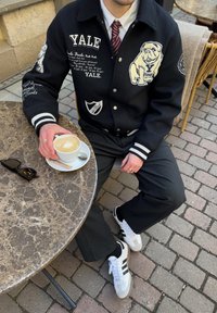 Casaco varsity preto com a marca Yale e emblemas, calças cinzas, sapatilhas brancas, segurando um cappuccino numa mesa de mármore.
