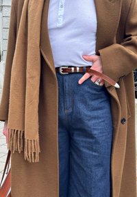 Personne portant un manteau marron et une écharpe, chemise blanche côtelée rentrée dans un jean bleu taille haute avec une ceinture en cuir marron, main dans la poche.