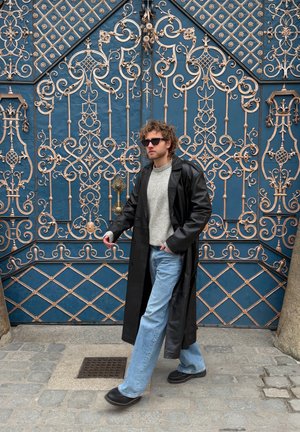 Hombre con abrigo de cuero negro, suéter gris, vaqueros azules y gafas de sol caminando por una calle adoquinada frente a unas rejas de hierro forjado azules y doradas ornamentadas.
