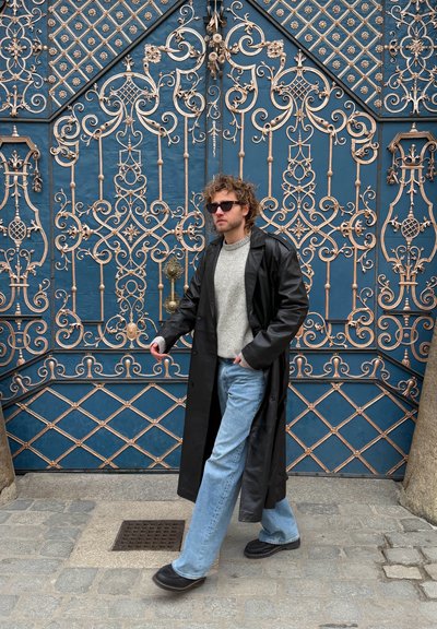 Hombre con abrigo de cuero negro, suéter gris, vaqueros azules y gafas de sol caminando por una calle adoquinada frente a unas rejas de hierro forjado azules y doradas ornamentadas.