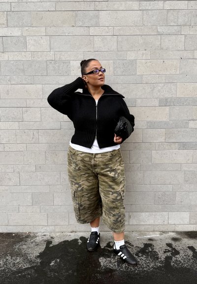 Chaqueta negra acanalada con cremallera, camiseta blanca, pantalones cortos cargo de camuflaje y zapatillas negras. Accesorios incluyen gafas azules y un bolso de mano. Fondo de ladrillo gris.