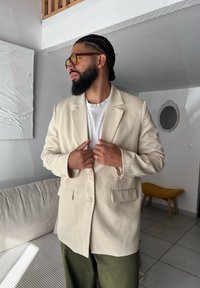 Ανοιχτό μπεζ σακάκι με ανασηκωμένους γιακάδες, δύο μπροστινές τσέπες και κλείσιμο με τρία κουμπιά, που φοριέται πάνω από λευκό πουκάμισο, με ελαιόχρωμες παντελόνες.