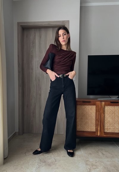 Top burdeos de manga larga con textura fruncida, jeans negros de cintura alta y bailarinas negras. Bolsa negra minimalista y armario de madera al fondo.