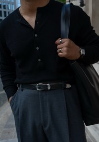 Camisola henley preta com nervuras e pala de botões, calças cinzentas plissadas, cinto de pele preta com fivela prateada, bolsa de pele preta.