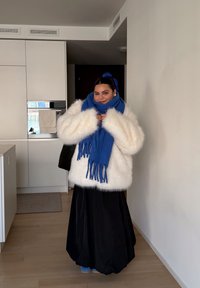 Fehér bolyhos kabát, kék rojtos sál, fekete légies szoknya, kék lábbeli. Letisztult konyhai háttér világos fa padlóval.