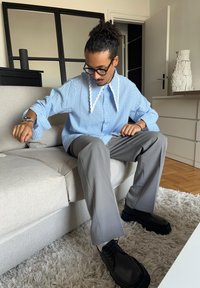 Blå gingham ternet langærmet skjorte med scalloped krave, grå bukser og sorte snørebåndssko, siddende på en lys grå sofa.