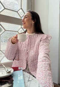 Quilted jakke i pink med blomsterprint, med flæsekorte ærmer og bånd, holdt af en person med en kop. Marmorbord og magasin synligt.