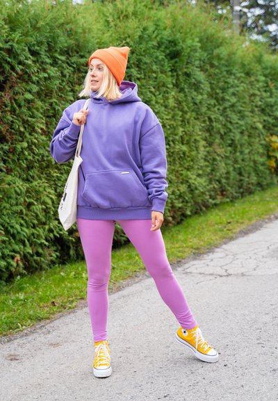 Sudadera grande morada, leggins rosas y zapatillas amarillas. Gorro naranja y bolsa de lona. Tejido texturizado, corte relajado y colores vibrantes.