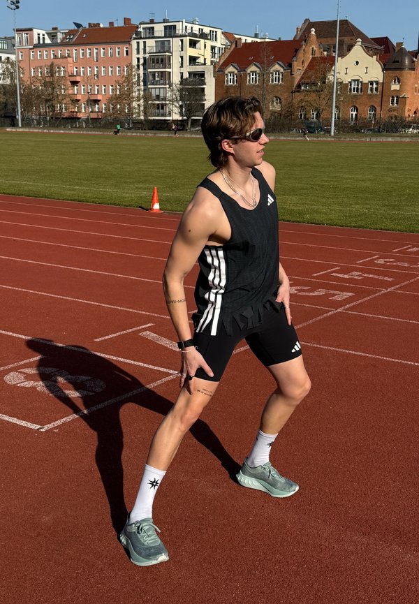 Runner wearing black athletic wear and sunglasses stretching legs on red track near grassy field and residential buildings.