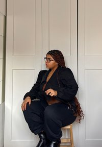 Μαύρο oversized μπουφάν με φουσκωτά μανίκια, συνδυασμένο με μαύρα παντελόνια και μπότες. Πλεκτά μαλλιά και στρογγυλά γυαλιά, καθισμένα σε μια ξύλινη σκαμνί.