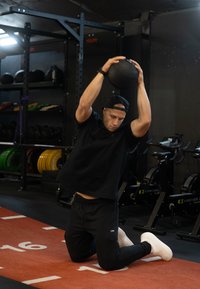 Čierne atletické tričko; čierne nohavice; biele tenisky; muž kľačiaci, zdvíhajúci čiernu medicinbal s textúrou nad hlavou; pozadie telocvične.