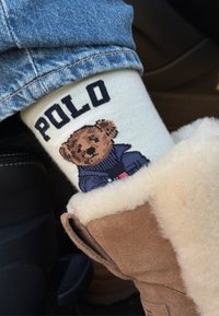 Kokvilnas zeķe ar krēmkrāsas pamatu, uz kuras attēlots lācis zilā džemperī un melnā krāsā uzraksts "POLO". Saskaņota ar brūnu šērlinga zābaku.