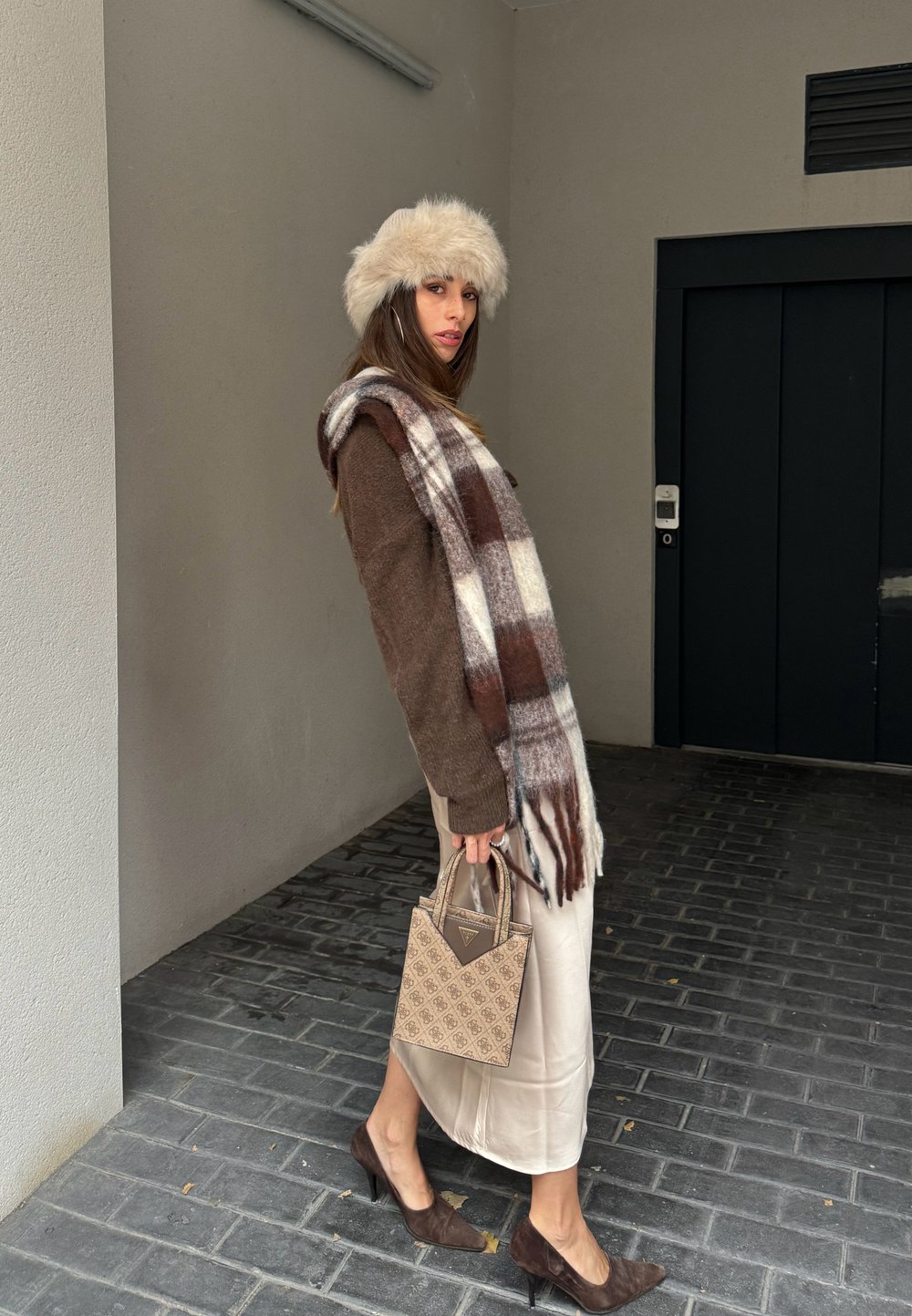 Brown wool jumper, plaid scarf featuring white and brown tones, beige satin skirt, brown suede heels, and a textured beige handbag.