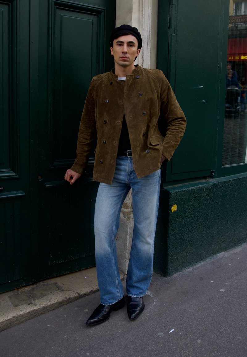 Homme portant une veste marron à double boutonnage, une chemise noire, un jean bleu clair et des chaussures noires, appuyé contre des portes vert foncé d'un bâtiment sur un trottoir en ville.