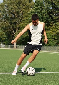 Idrettsutøver i en ermeløs hvit og svart drakt med gullakenter, svarte shorts og rødsålede fotballsko, dribler en svart-hvitt fotball på gress.