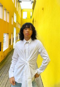 Witte langemouwen shirt met een twistdesign aan de voorkant, aangevuld met lichte denim jeans. De achtergrond heeft gele muren en een houten vloer.